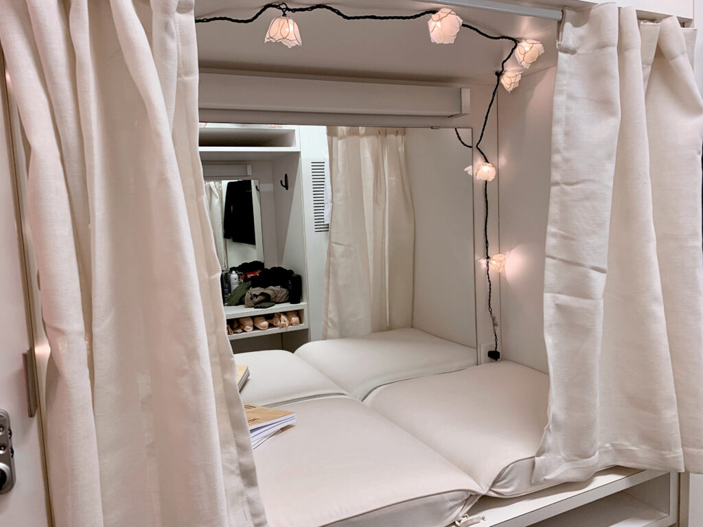 The photo shows a dressing room mirror and tabletop that's been transformed into a curtained cubby with twinkle lights strung up inside. The short, beige curtains are attached to a pressure rod, and a matching padded mat is lain across the top of the table surface to provide a place to sit or curl up with a book.