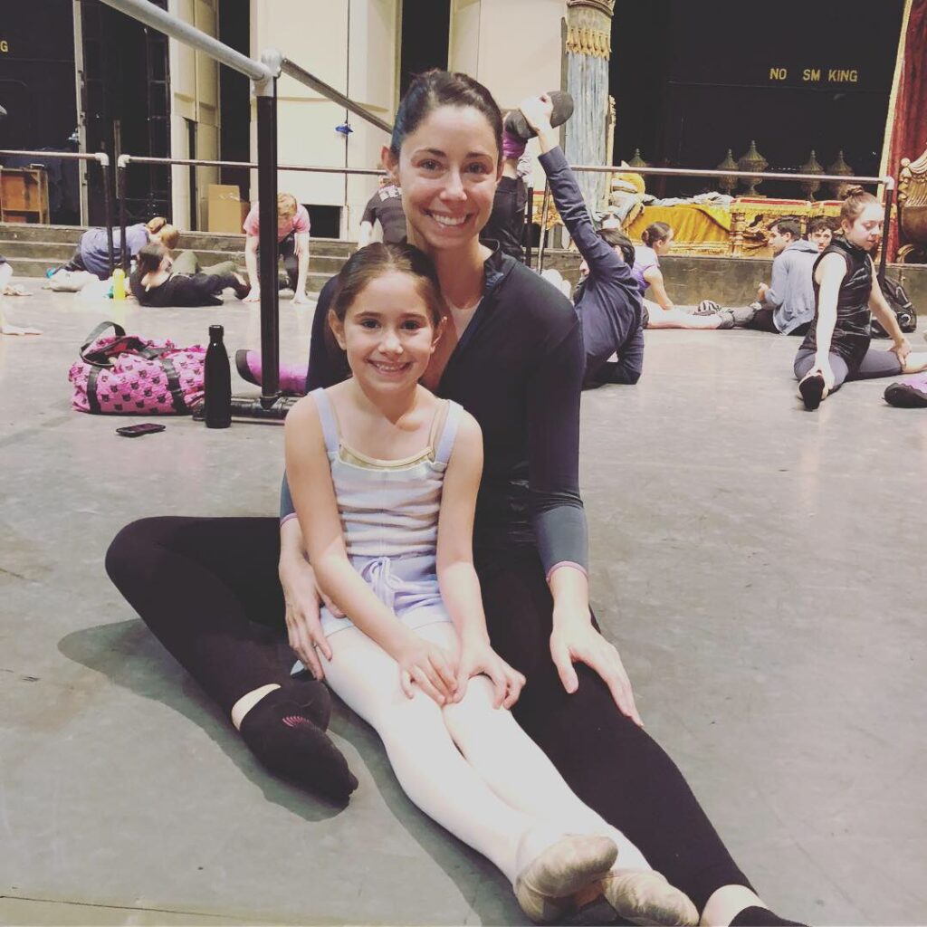 Sasha Vincett, wearing a dark, long-sleeved leotard, black tights and black socks, sits on the stage floor next to a portable ballet barre. Her young daughter, wearing a lavender leotard, pink tights and pink slippers, sits in between Vincett's legs, with her own legs out straight in front of her. They smile for the camera. Behind them, other dancers in workout clothing sit on the ground and stretch.