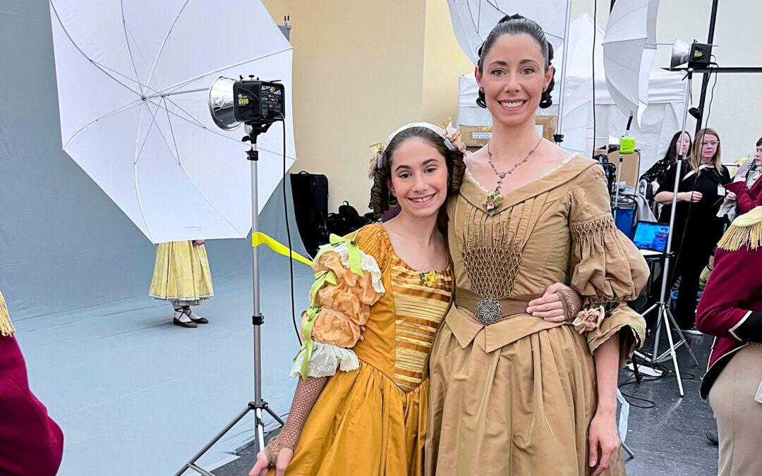Sasha Vincett and her young daughter wrap their arms around each other's waist and smile for a picture. They wear Victorian-era dresses in shades of gold and light brown, with their hair pulled back and in ringlets. Behind them, a photo shoot is happening with other dancers in costume.