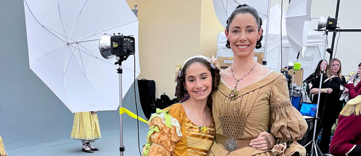 Sasha Vincett and her young daughter wrap their arms around each other's waist and smile for a picture. They wear Victorian-era dresses in shades of gold and light brown, with their hair pulled back and in ringlets. Behind them, a photo shoot is happening with other dancers in costume.