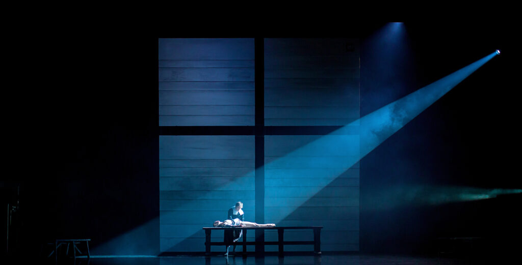 A zoomed-out photo shows a large cross and ghost light projection onstage. A man, dressed in Puritan clothing, leans over a long table where a woman lies prone. The light shines on them from the upper right diagonal.