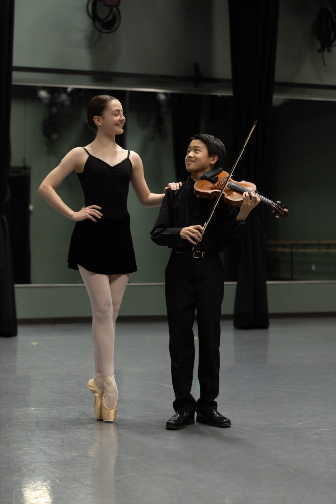 In a dance studio, a female ballet student stands in sous-sus on pointe in a black camisole leotard, a black pointe skirt, and pink tights. She smiles and rests her hand on the shoulder of a young male violin student, who stands to her right and looks up at her smiling, too. He wears all black formalwear and holds his violin and bow.