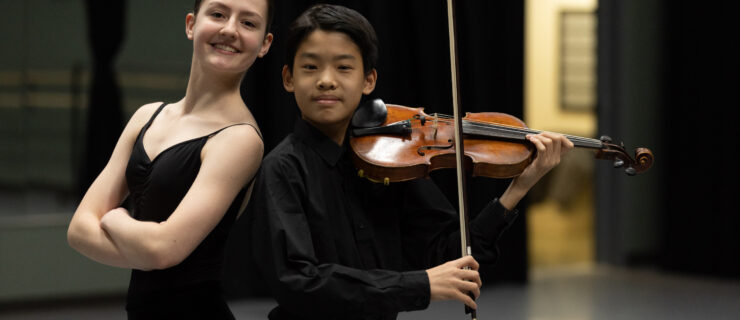 In a dance studio, a female ballet student and a young male violin student stand back-to-back, posing for the camera.