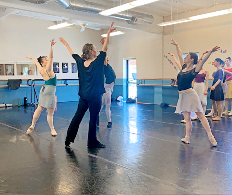 In a large dance studio, Julia Adam leads a rehearsal for "A Midsummer Night's Dream." Both male and female dancers rehearse. Adam wears flowy black clothing, and they all pose in tendu derriere in a circle, arms lifted in a V.