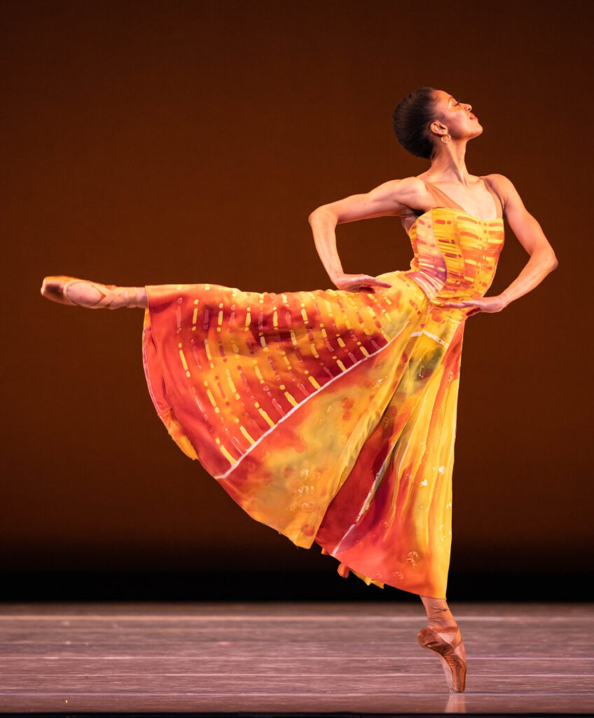Katlyn Addison does a piqué arabesque onstage, hands on hips with her chin lifted confidently. She wears a long, flowing yellow–red watercolor dress and pointe shoes.