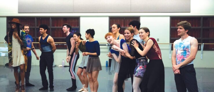 A group of ballet dancers in different rehearsal attire stand together in a line and laugh, clapping and looking around. Katlyn Addison wears a sundress and stands to the far left, smiling as she leads rehearsal.