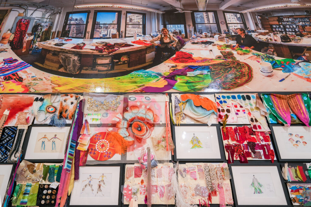 A wall of fabric swatches and costume illustrations, with a wall-width image of Willa Kim in a costume studio above the swatches.