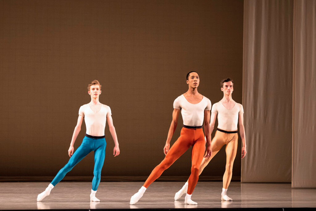 Loren Walton is in tendu plié in a parallel second position. He is onstage and wearing rust-colored tights, a black belt, white t-shirt, and white socks with white flat shoes.
