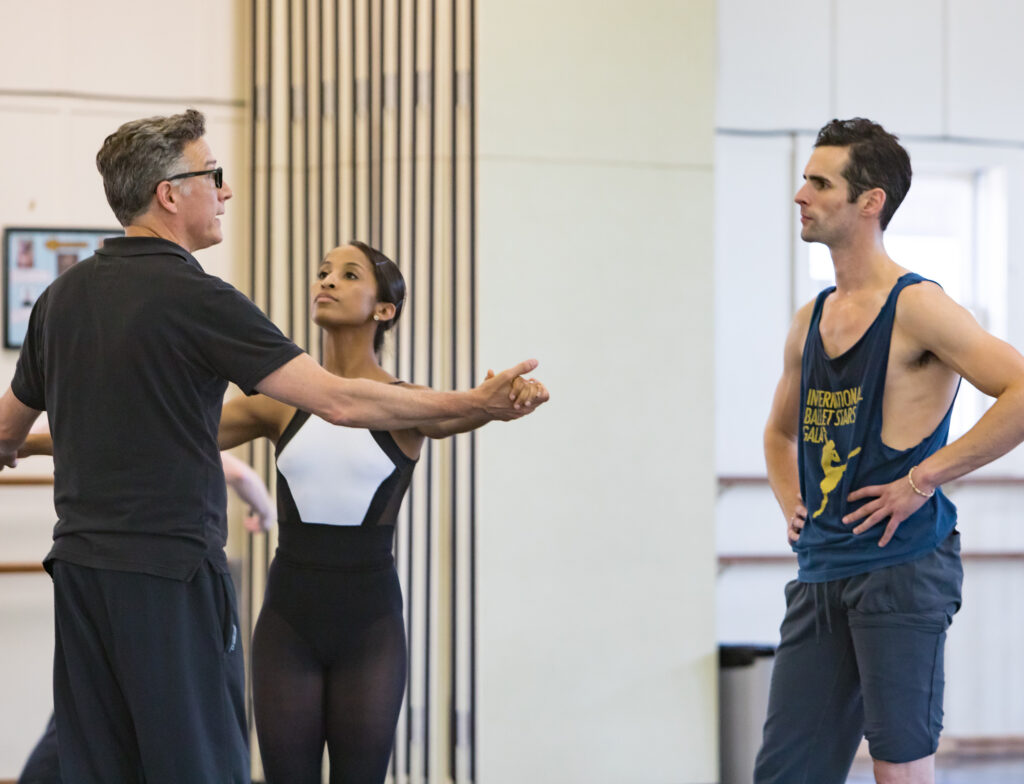 In a large dance studio, Paul Vasterling stands with his back facing the camera, wearing a black polo shirt and dark sweatpants. He joins hands with ballerina Kayla Rowser and holds their arms wide, as if dancing together. ROwser, wearing a black and white leotard and black tights, looks up at Vasterling as he talks. Owen Thorne, wearing a blue tank top and sweatpants, stands to the right of them, watching and listening intently.