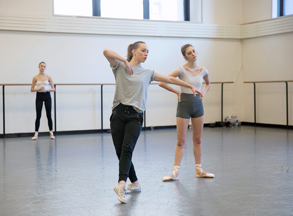 Adriana Pierce, wearing a gray T-shirt, black jeans and white Converse sneakers, demonstrates a dance move to a teenage female dancer. The young dancer—wearing gray shorts, a white leotard and pointe shoes—stands with her hands on her hips, watching Pierce in the mirror. Another teenage ballerina, in a white leotard, black tights and pointe shoes, stands in the back of the studio and watches, as well.