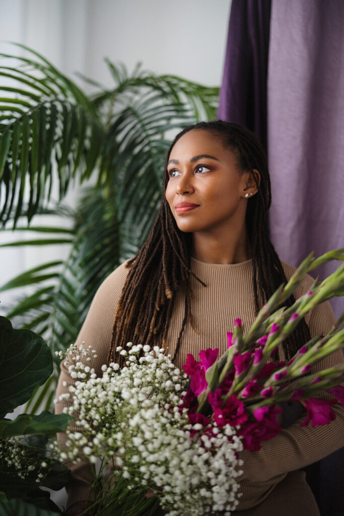 A headshot of "Dances" author Nicole Cuffy. She is wearing a brown, long-sleeved top and looking toward her right. Flowers are in the foreground of the photo. 