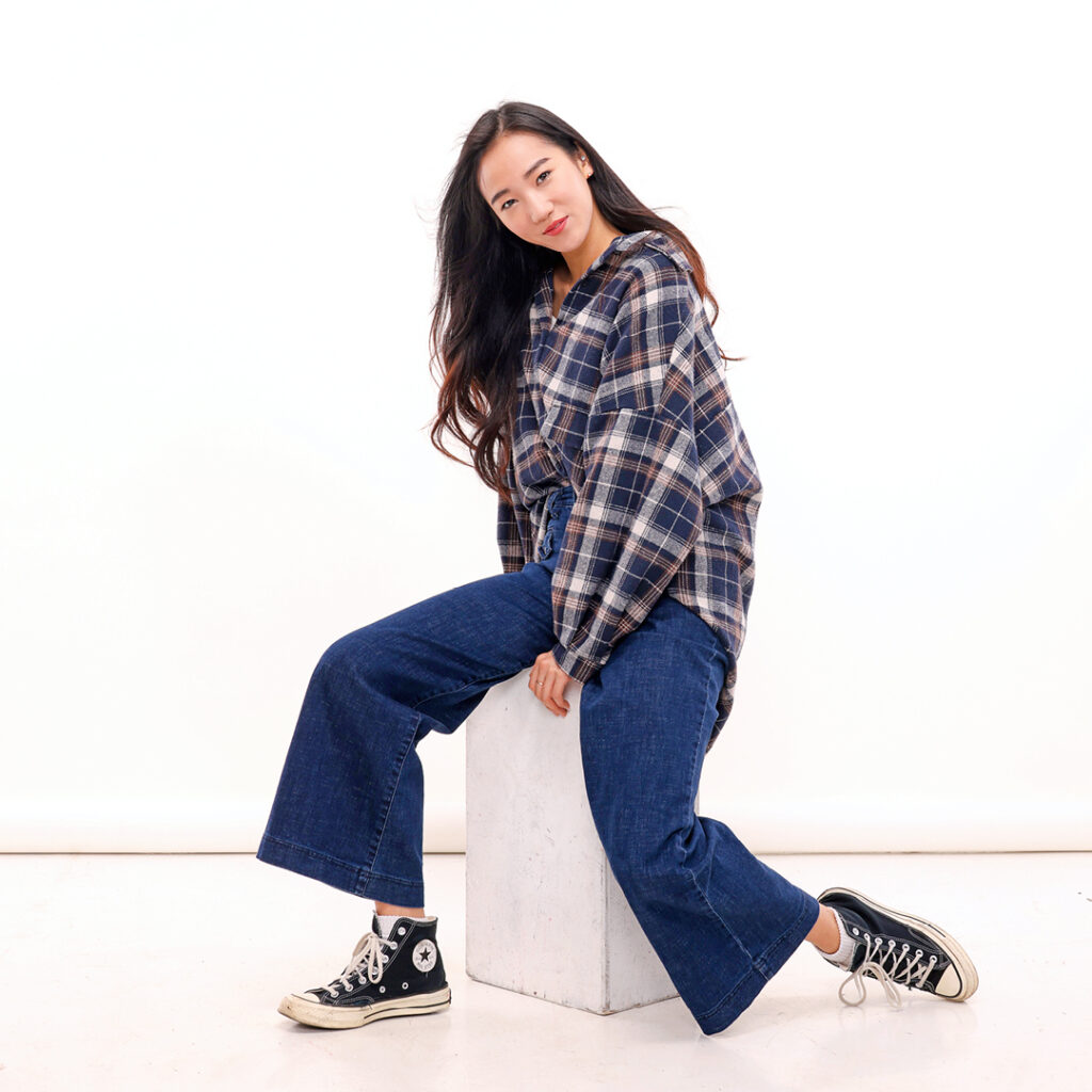 SunMi Park sits on a tall white box, wearing a large flannel shirt, bell-bottom jeans and black high tops sneakers. She twists her body slightly to her right, tucking her left leg behind her slightly and pointing her left foot. She holds onto the box with her left hand, cocks her head slightly to the right and looks directly toward the camera with a small smile.