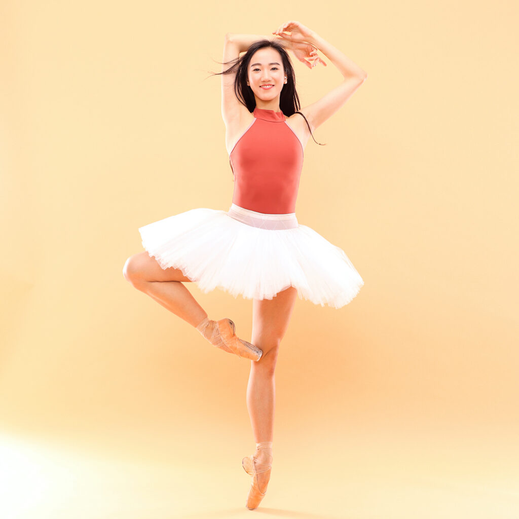 SunMi Park poses in retiré on pointe in front of a yellow backdrop. She stands on her left foot and raises her arms up, bending her right arm along the top of her head and her left arm out a bit, bending her elbow to create the same shape as her right leg in retiré. She wears a coral-colored leotard and white practice tutu, and smiles serenely for the camera.