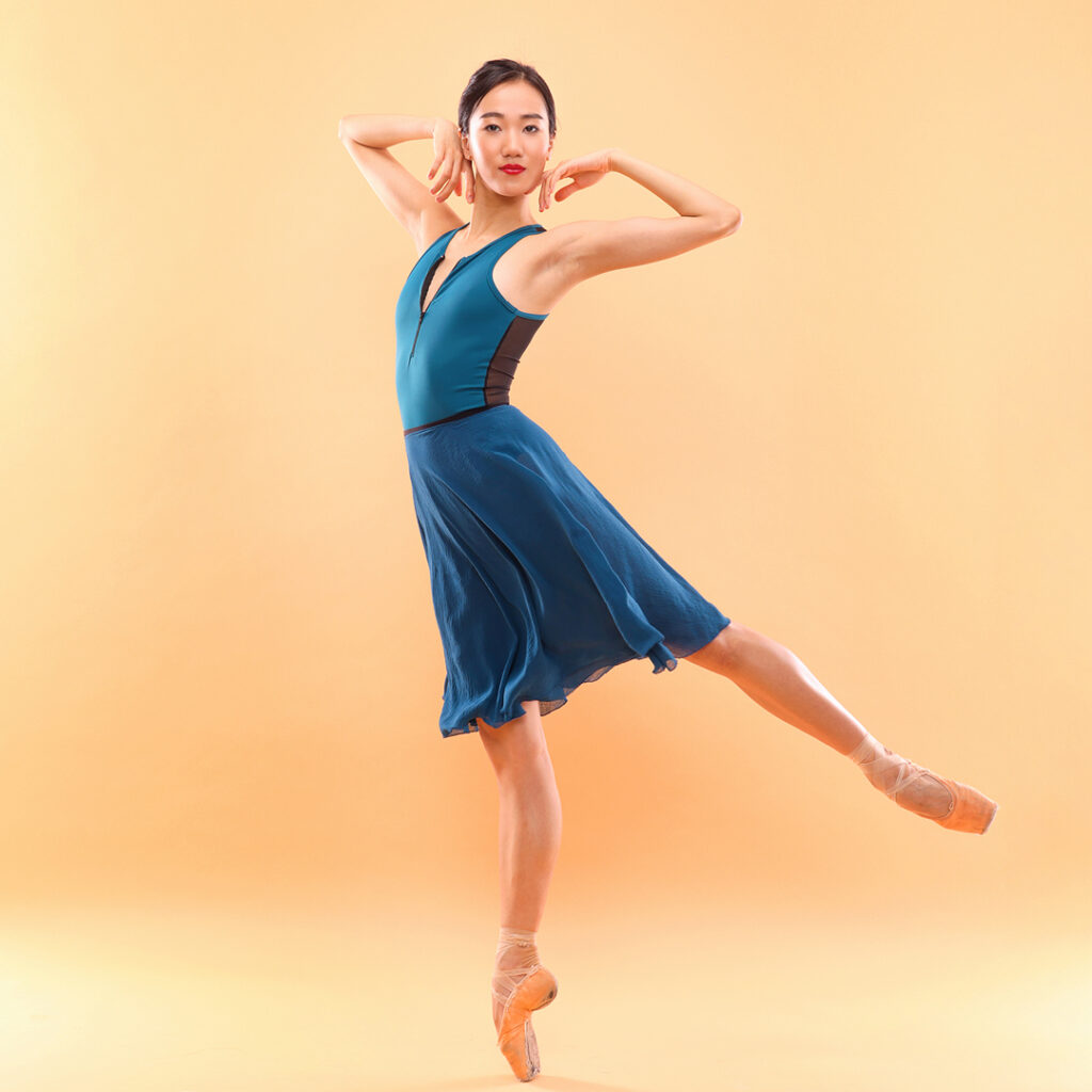 SunMi Park, wearing a turquoise leotard and matching knee-length skirt, poses on pointe in a low arabesque with her left leg raised. She frames her face with the backs of her hands, her amrs bent at the elbows. She looks directly front with a smart, closed-mouth smile. Her hair is pulled back into a bun.