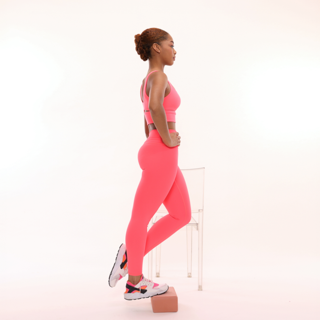 Model Azrielle Smith demonstrates the exercises as described. She is wearing pink leggings, a pink workout top, and pink, white, and black sneakers. She is using a pink yoga block and a chair.