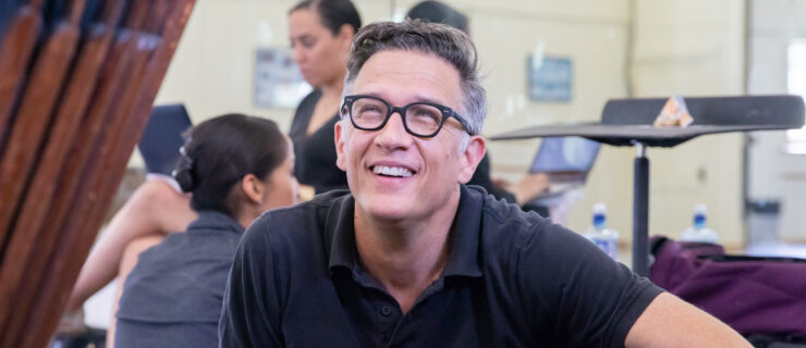 Paul Vasterling, wearing a black polo shirt and black-frame glasses, sits on the floor of a dance studio and looks up towards someone, smiling. Behind him another dancer sits facing the mirror, while an woman on the artistic staff sits crossed-legged in a chair, her back to the mirror, and looks at the laptop in her lap.