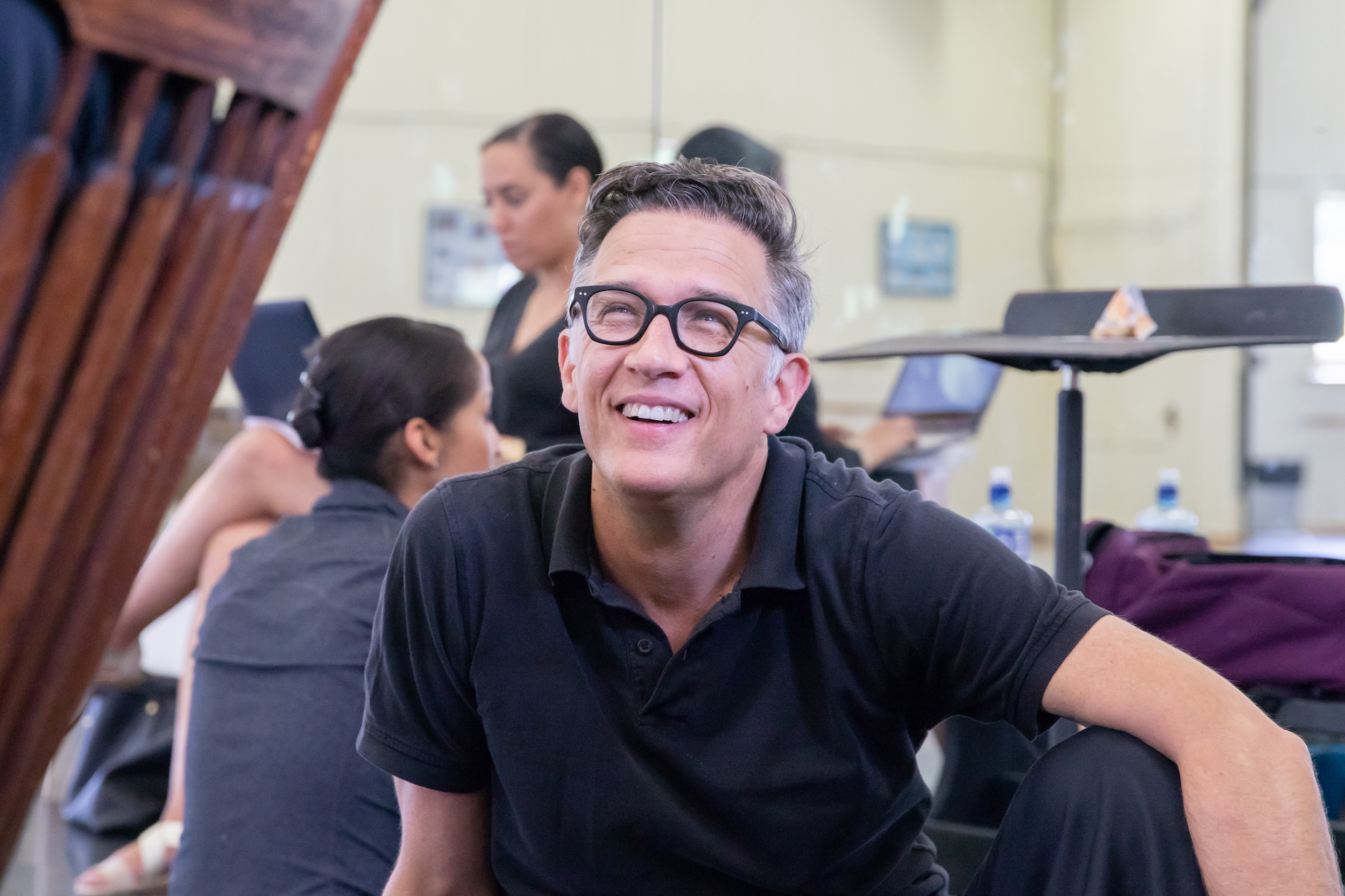 Paul Vasterling, wearing a black polo shirt and black-frame glasses, sits on the floor of a dance studio and looks up towards someone, smiling. Behind him another dancer sits facing the mirror, while an woman on the artistic staff sits crossed-legged in a chair, her back to the mirror, and looks at the laptop in her lap.