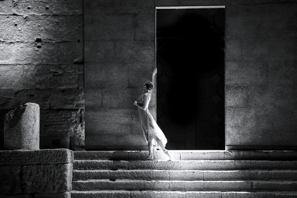 In this black and white photo, ballerina Anna Blackwell stands barefoot on a wide, shallow stone staircase, reaching up to touch the side of an entrance in the stone wall. She wears a filmy dress and looks up at the wall, the back of her head to the audience. She stands with her legs in parallel, her left foot reaching back as she walks.