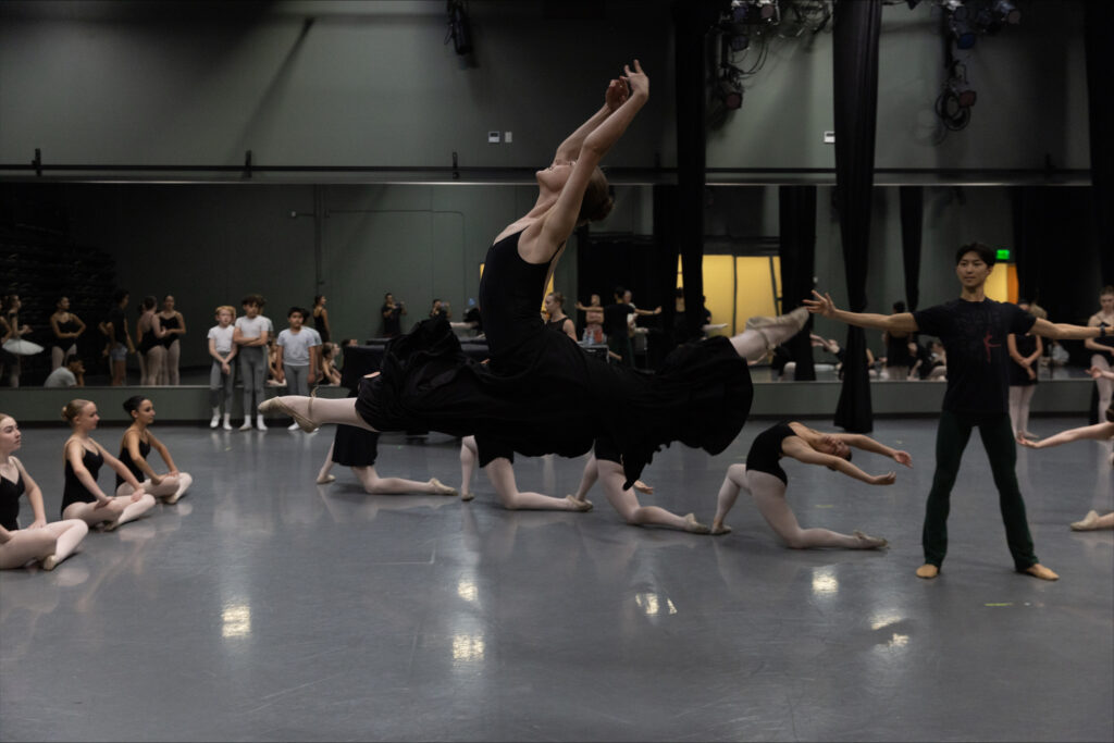 In a large dance studio, School of Ballet Arizona students rehearse for "Raymonda" in black practice clothes. In the middle, the lead female principal does a flying grand jete, and younger students pose on their knees in a semi-circle around her.