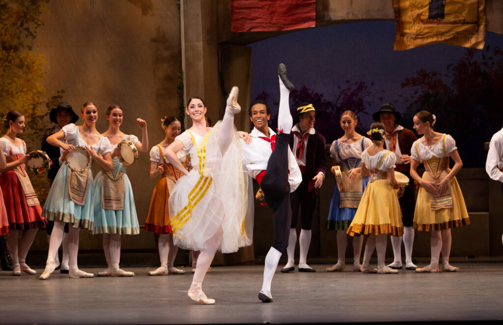 Sasha Vincett and Ricardo Santos lock arms and kick their right legs high in the air during a performance. They wear huge smiles on their faces. Vincett is costumed in a white peasant dress with yellow trim and a tulle skirt. Santos wears a white peasant blouse with a red tie and cummerbund, black knickers, white tights and black ballet slippers. Behind them a large group of dancers wearing peasant costumes look on, with some holding tambourines.