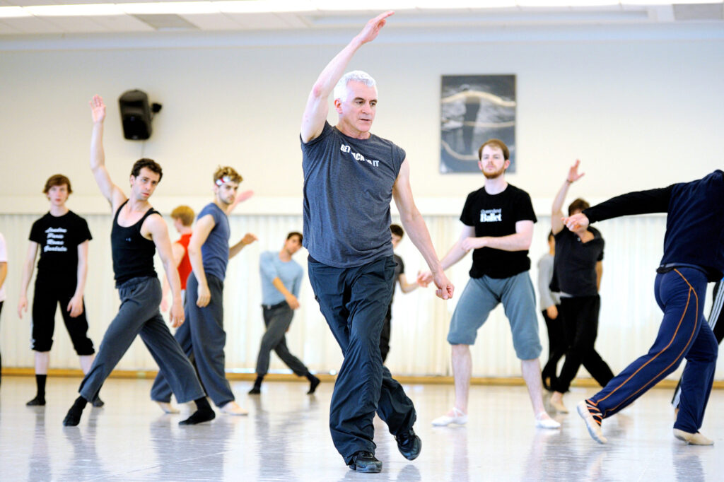 In a bright studio, Val Caniparoli leads a rehearsal filled with SFB dancers in various iterations of athletic dancewear. Caniparoli wears a grey tshirt and black pants.