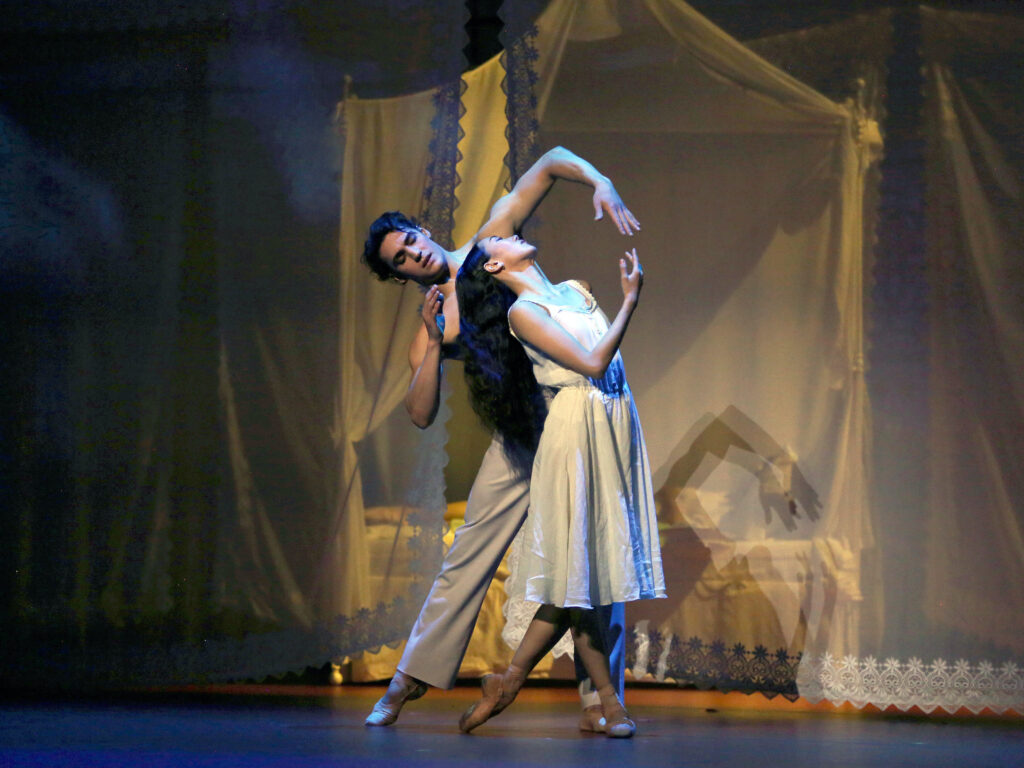 SunMi Park and Daniel Camargo dance a pas de deux onstage during a performance. Park, wearing a knee-length white dress, stands in profile with her left foot in B plus, leaning her upper body back and bending her right arm so that her right hand almost meets Camargo's left hand, which reaches over her. Camargo, shirtless and in white pants, stands on his left leg and extends his right leg out, and lightly caresses Park's long, dark hair with his right hand. Behind them is a bed draped in filmy white curtains.