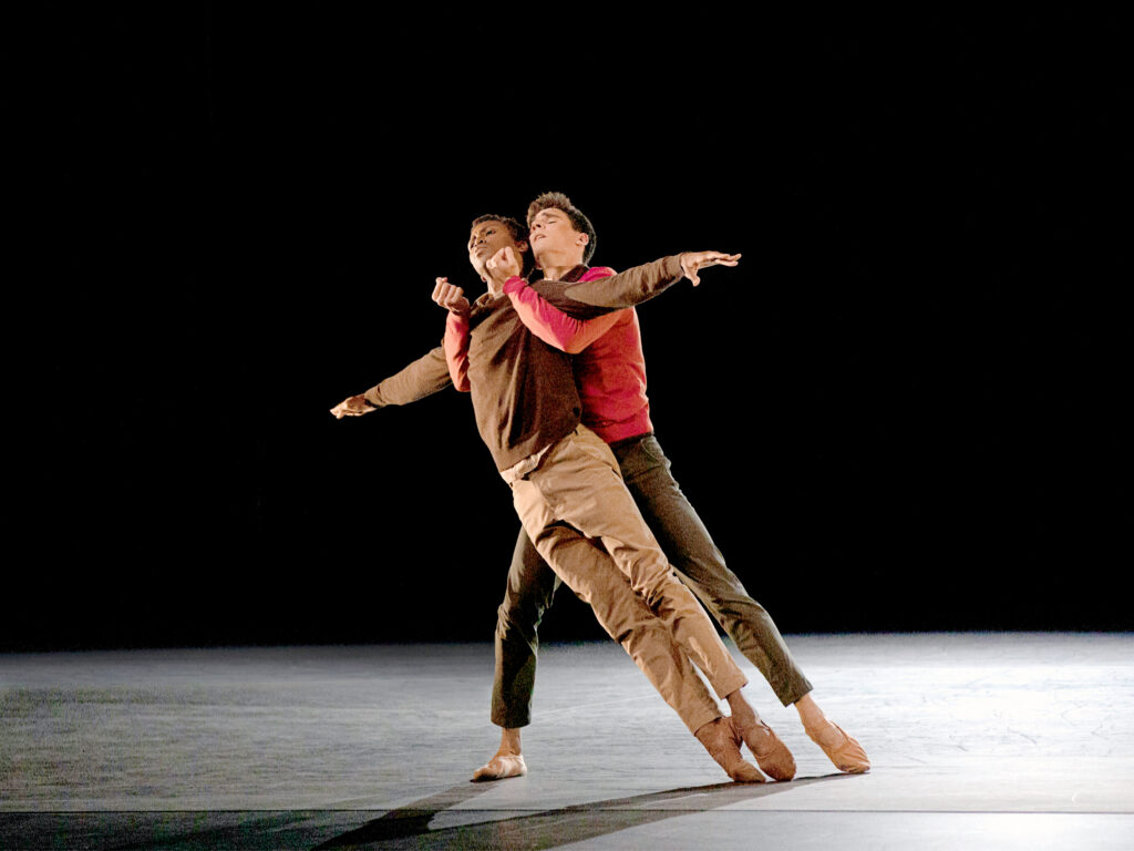 João Menegussi lunges slightly to his right leg, holding Calvin Royal III underneath the armpits with his arms during a romantic pas de deux. Royal III leans with him, his arms stretched out to the side and his legs tight together in parallel, both feet pointed. They close their eyes. Both men wear chinos and solid-colored sweatshirts, and ballet slippers.