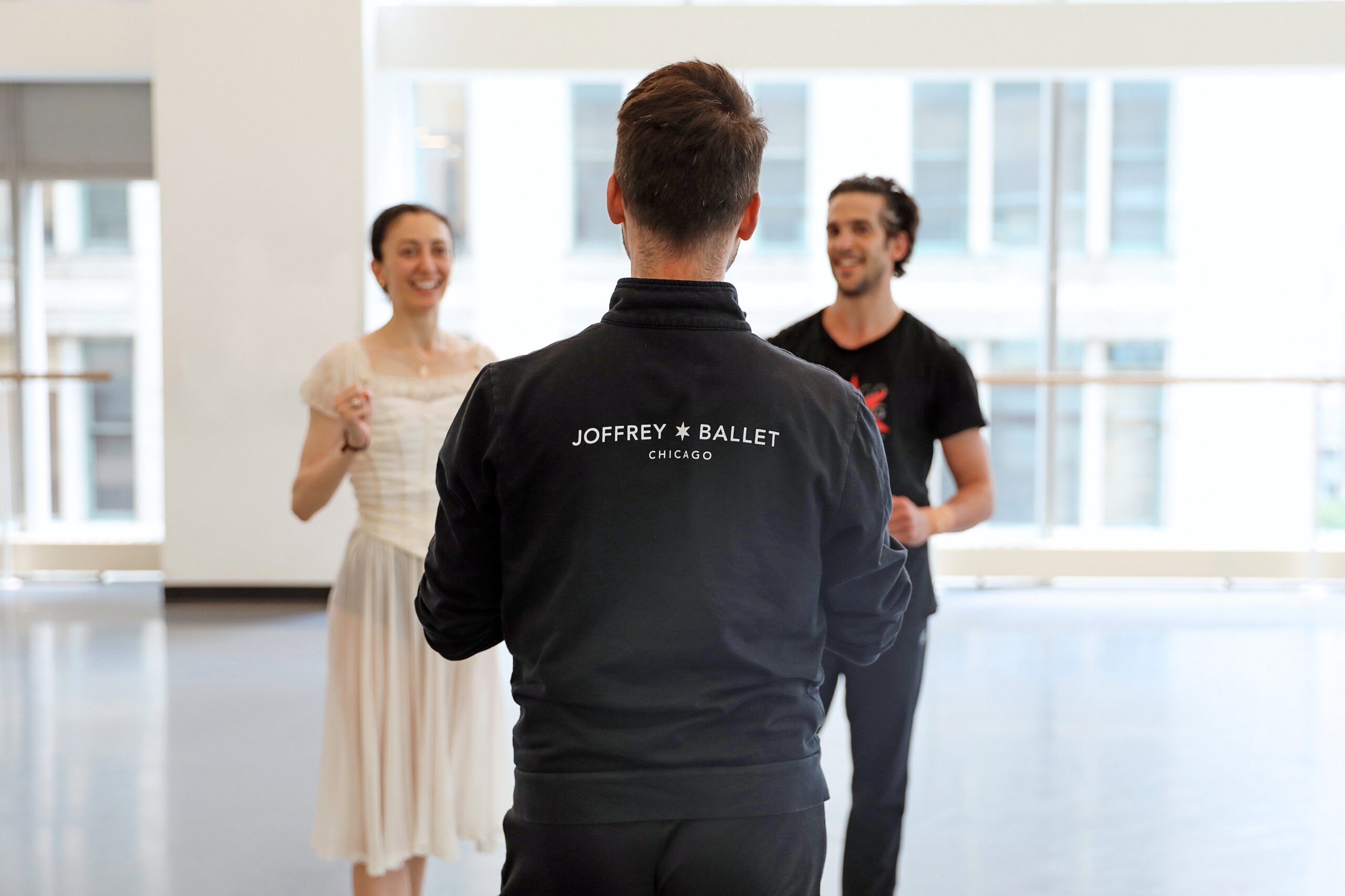 Adam Blyde is shown from behind in a large, airy dance studio, standing in front of dancers Victoria Jaiani and Alberto Velazquez, talking to them. He wears a black zip-up sweatshirt with the words "Joffrey Ballet Chicago" on the back. Jaiani wears a costume of a filmy white dress, while Velazquez wears a black T-shirt and black athletic pants; they both laugh as Blyde talks to them.