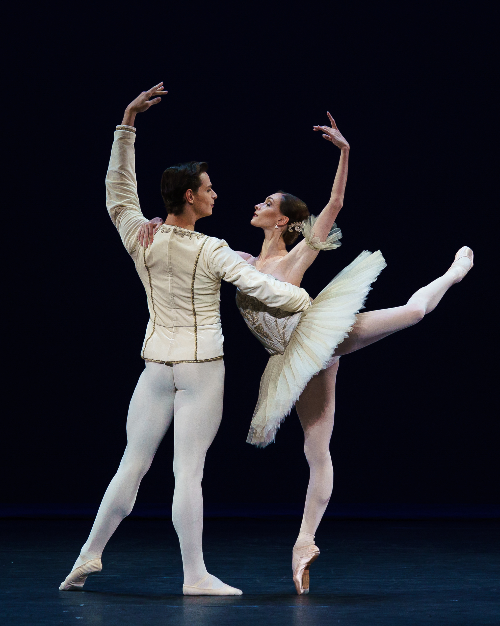 A man and a woman perform the 0 pas de deux onstage, wearing cream and gold bejeweled costumes in the classical ballet style.