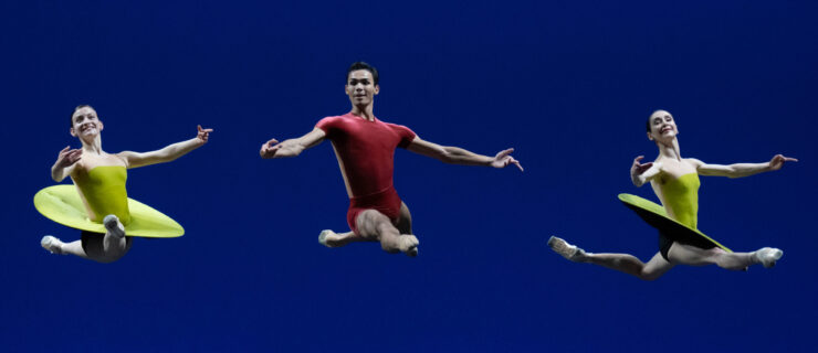 In front of a deep blue background, two female and one male dancer do a Bournonville jeté en face toward the audience. He wears a red short-sleeve biketard, and they wear bright green modern pancake tutus.
