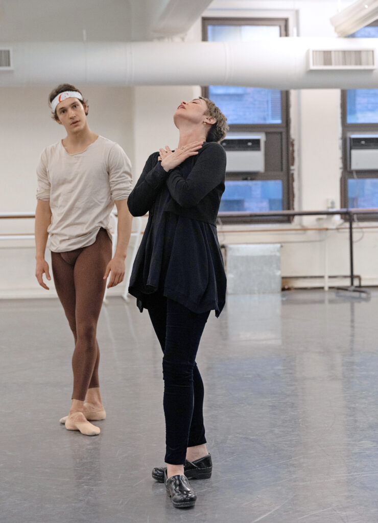 Susan Jaffe, wearing a long black shirt, black leggings, and clogs, crosses her hands at her throat and looks up at the ceiling as she demonstrates a movement to dancer Daniel Camargo. He stands behind her to her right and watches intently. He wears a beige long-sleeved T-shirt, brown tights and tan ballet slippers, and has a thin white bandana tied around her forehead. They work in a dance studio with white walls and air conditioning units in the windows.