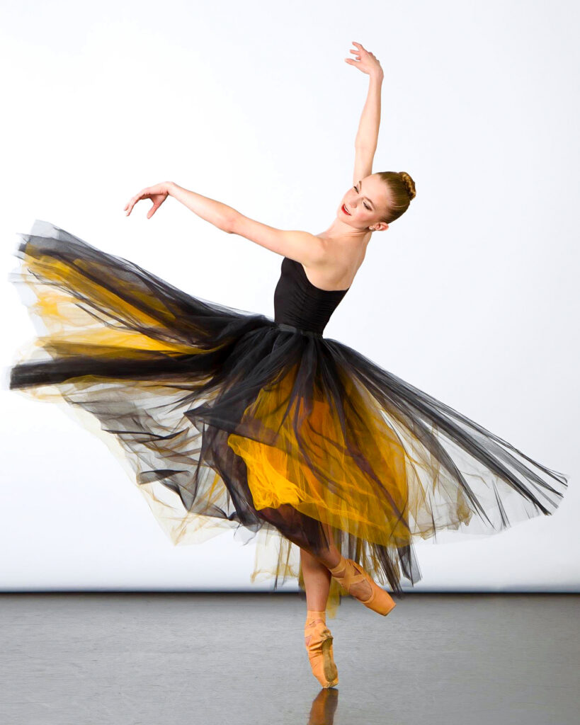 Photo of ballet dancer Sarah Whitehead on pointe with her left foot in coupé derriere and her arms in fourth arabesque. Her body faces upstage right and she looks back over her left shoulder. She wears a long, dramatic tutu with a black bodice and black and yellow tulle skirt.