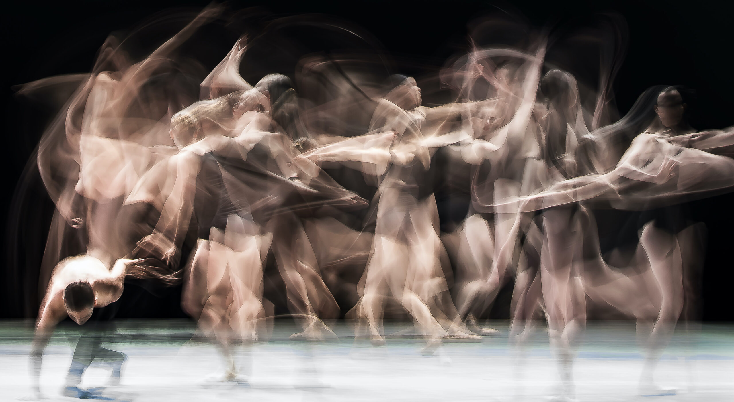 A landscape photo shows a group of dancers in motion onstage, their bodies blurred as they dance. They wear black leotards and shorts.