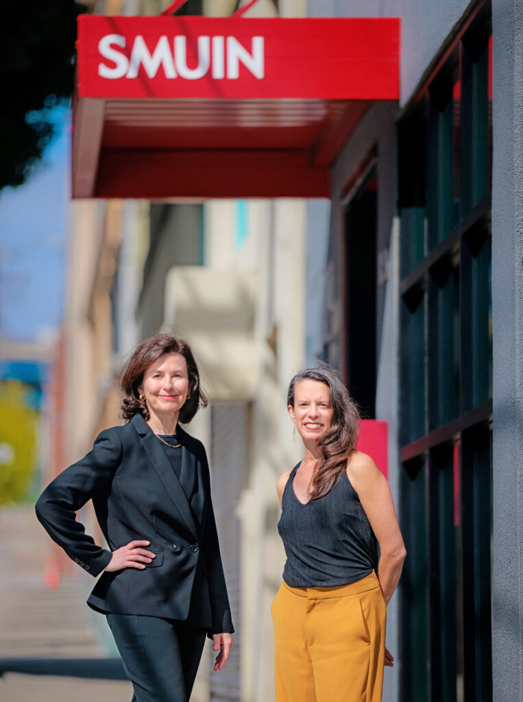 Celia Fushille, wearing a dark pantsuit, and Amy Seiwert, in a dark sleeveless top and yellow pants, stand side-by-side outside on a San Francisco street. Above them is a red sign that reads Smuin in white capital letters.