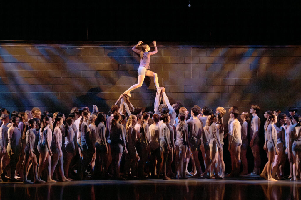 A large group of young ballet students perform onstage in a high-energy ensemble piece, wearing brown, black, and yellow costumes.