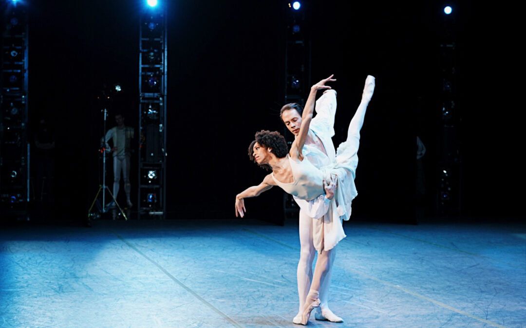 Chyrstyn Fentroy and Paul Craig are photographed from the wings dancing onstage during a performance of Chaconne. Fentroy does a penché arabesque on pointe in croisé with her left leg lifted and her arms out to the side. Criag stands to the right of her and holds her around the waist in the crook of his right elbow, reaching his left arm bakc behind him. Fentroy look out to the audience and wears a white dance dress, pink tights and pink pointe shoes. Craig wears a white blouse open at the neckline with billowing sleeves and white tights and ballet slippers.