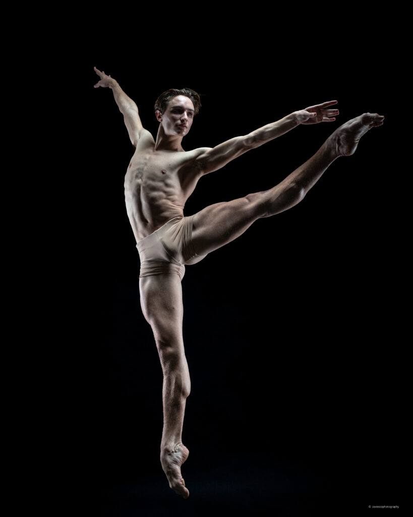 David O'Matz jumps with his right leg in an la seconde battement, his arms in an extended fourth. He looks over his pointed foot intently and is lit with a spotlight.