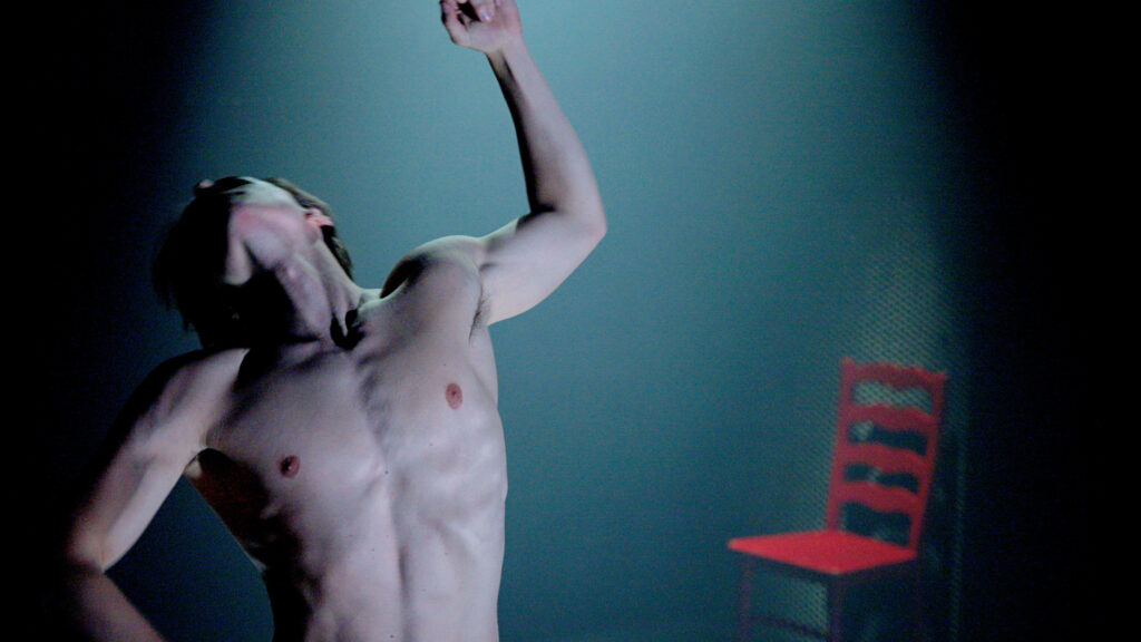 A close-up on Dylan Wald shows him stomach-up, moving toward the camera as he arches his chest back and flings his arms. He is lit by a spotlight, and a red wooden chair rests in the background.
