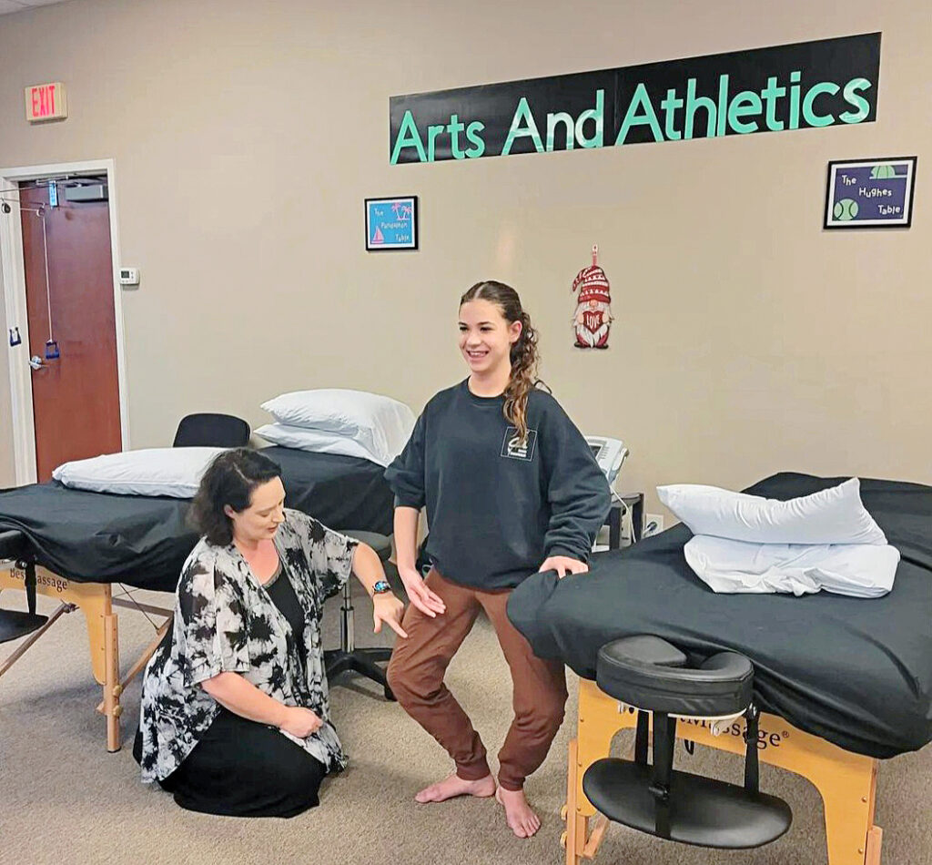 Emily Becker is shown kneeling on the floor of a physical therapy clinic, resting her left elbow on a stool and touching the knee of dancer Ally Francis, who is doing a plié in first position while holding a therapy table for support with her left hand. The dancer is barefoot and wears brown sweatpants and a black sweatshirt. Becker wears a black and white shirt and black pants.