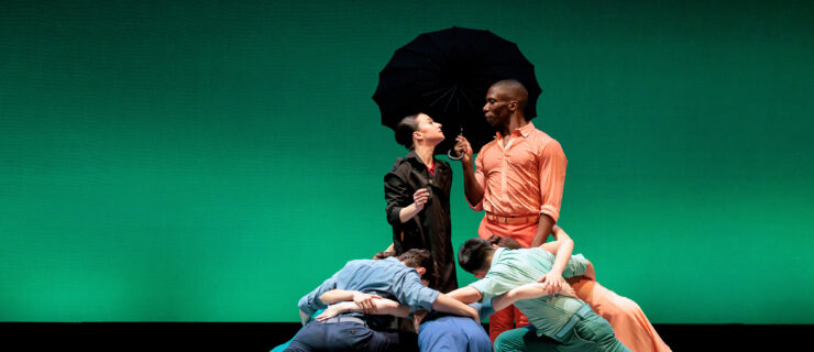Terez Dean Orr and Brandon Alexander stand face to face onstage in front of a green backdrop during a performance. They look wistfully into each others eyes. Alexander wears a pink shirt and pants and holds a black umbrella, while Dean Orr wears a dark rain coat. A group of danders in blue and green costumes encircle them, lunging deeply and with their arms around each others waists.