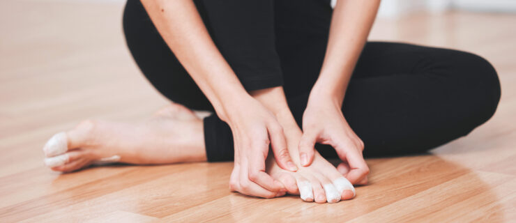 Shot of a young female ballet dancer sitting on the ground rubbing her injured foot. Her face is not shown. She wears a brown leotard and black tights and has athletic tape on several toes to protect them from blisters.