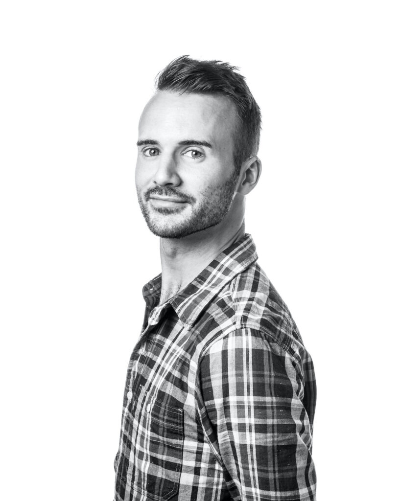 In this black and white portrait, Adam Blyde is shown from the ribs up, his body turned to the right as he looks over his left shoulder to smile towards the camera. He wears a plaid button down shirt.