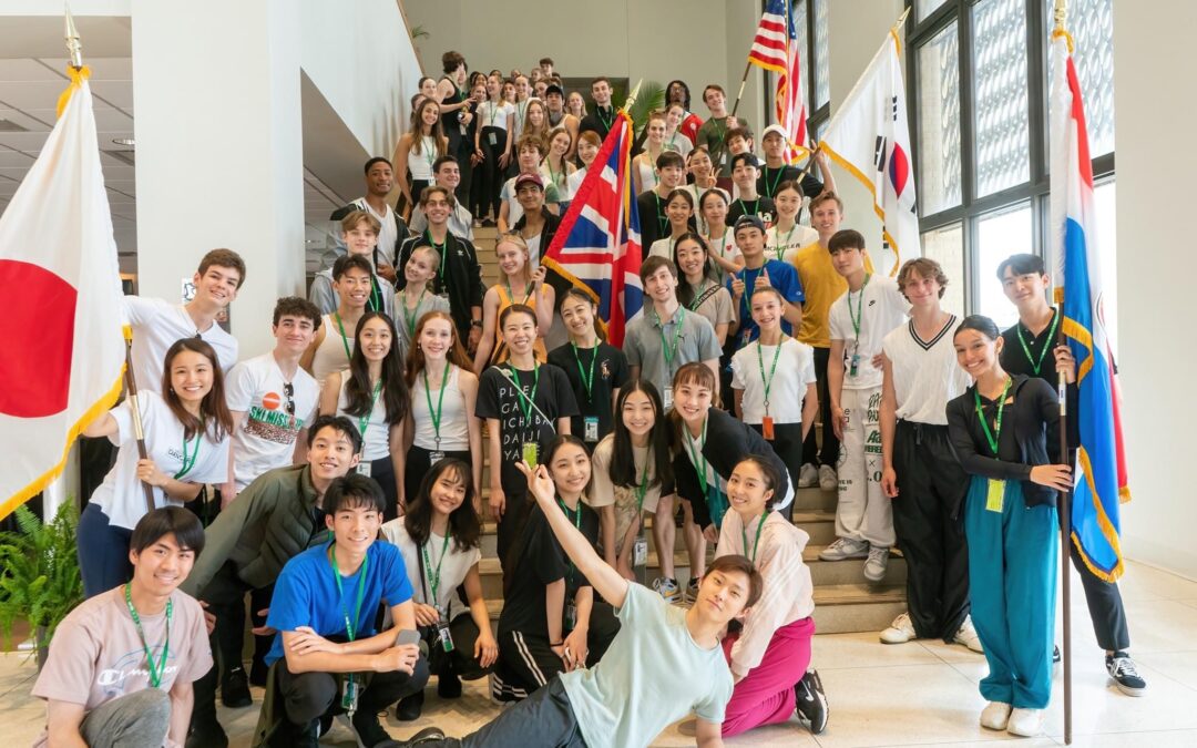 A large international group of dancers ranging between 15 and 27 years old gather on a marble staircase for a casual group photo. They wer either srteet clothing or athleisure wear, and five of them hold flags to various nations.