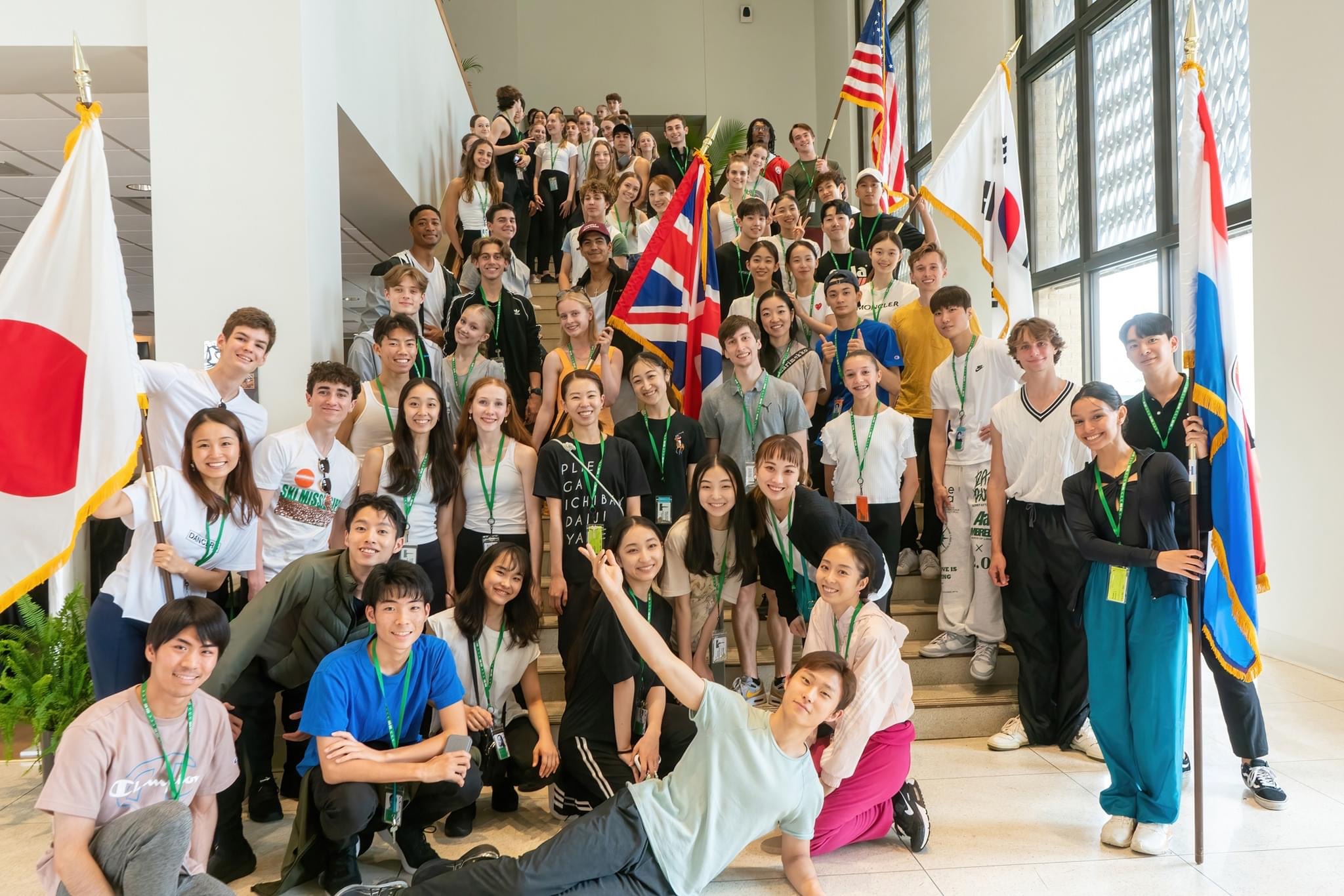 A large international group of dancers ranging between 15 and 27 years old gather on a marble staircase for a casual group photo. They wer either srteet clothing or athleisure wear, and five of them hold flags to various nations.