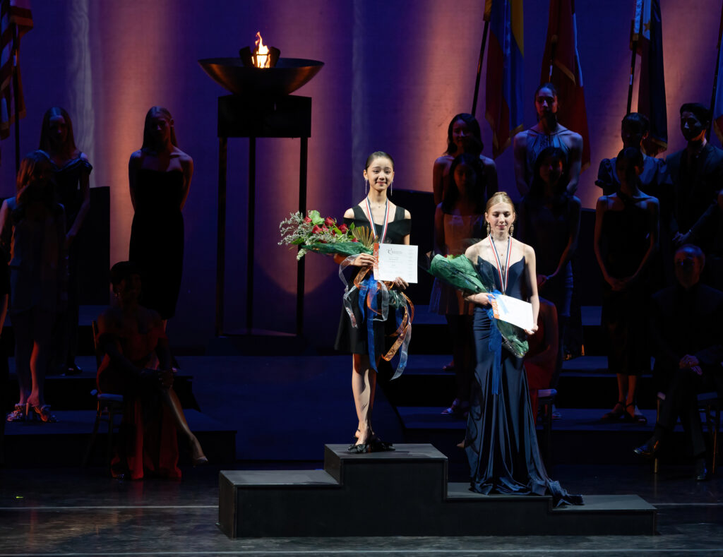 Two young women stand on an Olympic-style podium, wearing formal dresses and high heels and carrying bouquets of flowers. They each wear a medal around their neck.