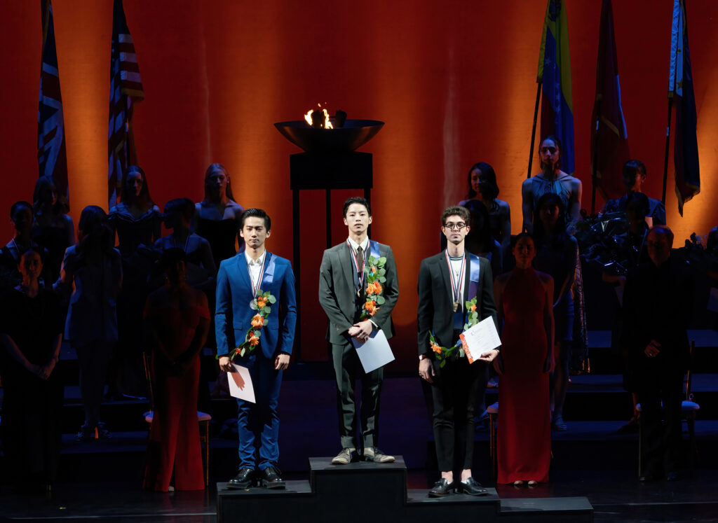 Three young men stand on an Olympic-style podium, wearing formal suits and a sash of flowers around their left shoulder. They each wear a medal around their neck.