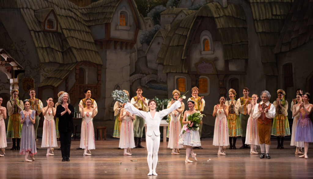 Ashton Roxander thanks the audience onstage after a performance of "Coppelia." He wears a white tunic and tights and extends his arms in a wide V overhead, smiling.