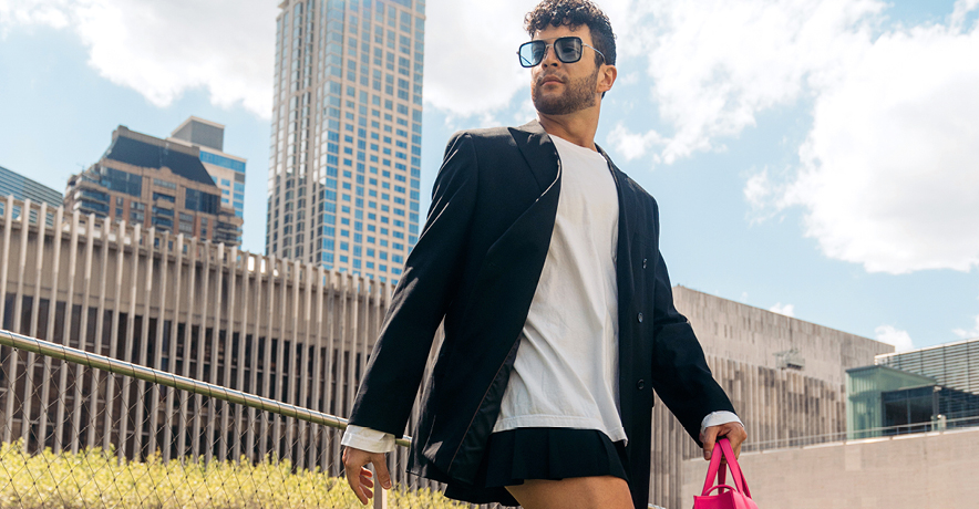 Gilbert Bolden III walks along a grassy knoll outdoors in New York City on a sunny day. He wears a black blazer, large white T-shirt, and black mini-skirt. He looks over his right shoulder and carries a hot-pink bag in his left hans. A large high-rise building looms in the background.