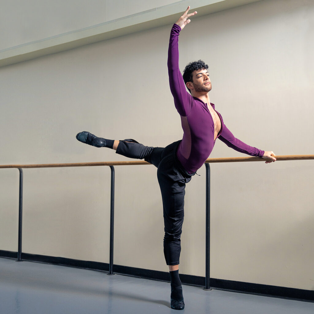 In a large dance studio, Gilbert Bolden III wears a burgundy, long-sleeved biketard with black warm-up pants, black socks and black leather ballet slippers. He poses at the barre in an attitude derriere in croisé with his left leg raised and left hand on the barre.
