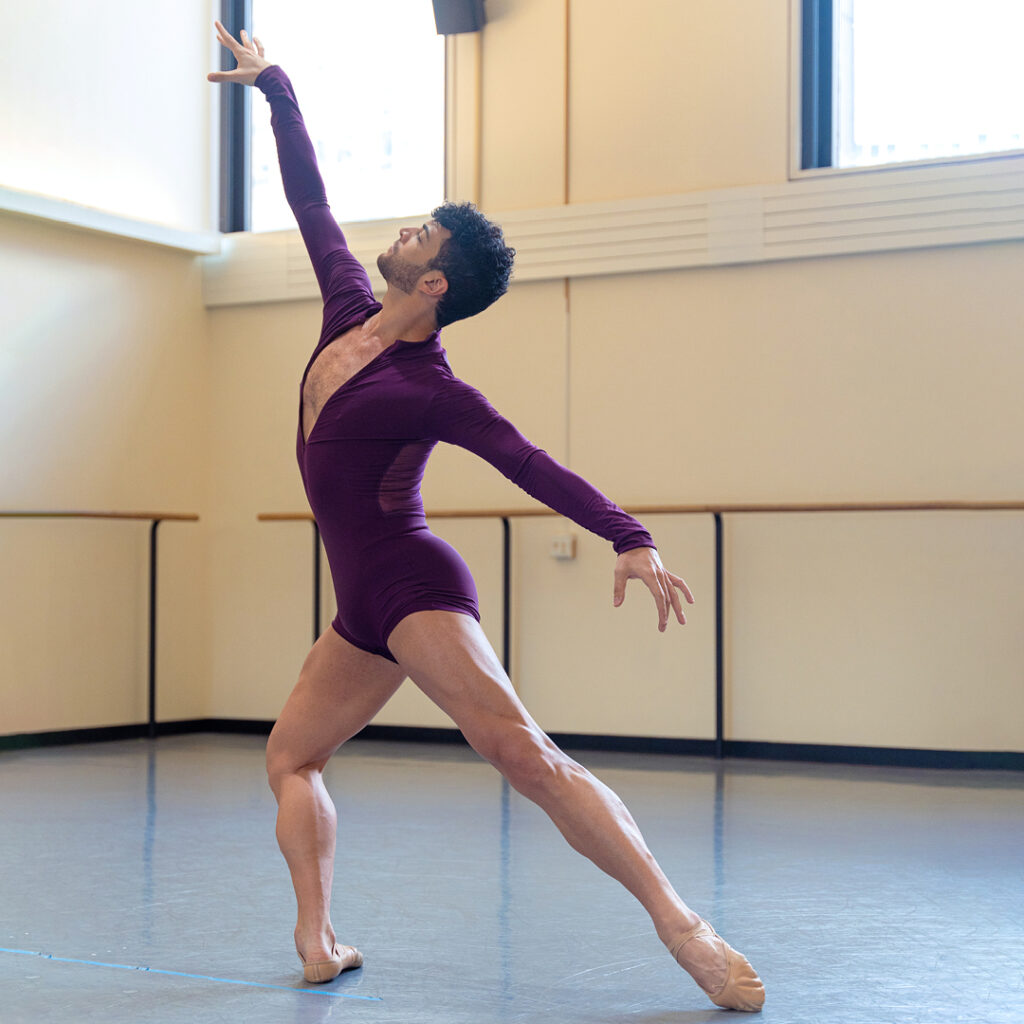 Gilbert Bolden III wears a burgundy, long-sleeved biketard and tan ballet slippers. He poses in a large dance studio with windows near the ceiling. He lunges to the right on his right leg, stretching his arms into a first arabesque line and pointing his left foot in tendu derriere.